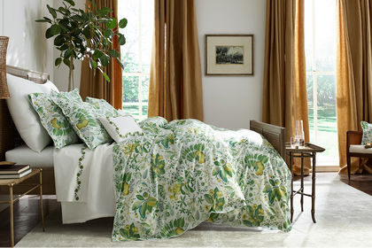 Master bed dressed in green and white floral duvet covers and pillow shams in a sunlit bedroom with golden drapes.