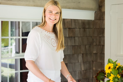 Portrait of Kate Schelter smiling in a white eyelet top in front of a wooden shingled wall.