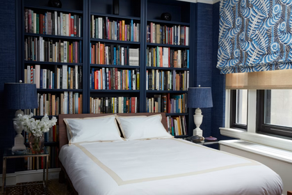 Bed styled with white bedding set in front of floor-to-ceiling dark blue built-in bookshelves filled with books.