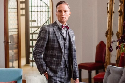 Designer Ken Fulk standing indoors in a plaid jacket surrounded by eclectic furniture and decor.