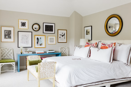 Wide view of a bedroom styled with white linens, coral pattern pillows, a round convex mirror above the bed, and a gallery wall behind a blue desk.