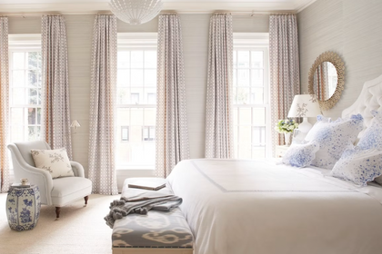 Bright primary bedroom featuring high windows with neutral patterned drapes, an upholstered king bed, and an armchair next to a blue-and-white garden stool.