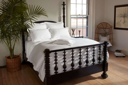 A neatly made bed with crisp white bedding and multiple white pillows, set against a tall tufted beige headboard in a softly lit bedroom. A dark bedside lamp and small decor items sit on a wooden nightstand to the left.