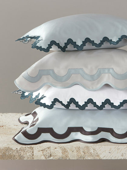Four stacked satin pillowcases in white and pale blue with scalloped edges and contrasting zigzag trim, displayed on a stone surface against a neutral background.