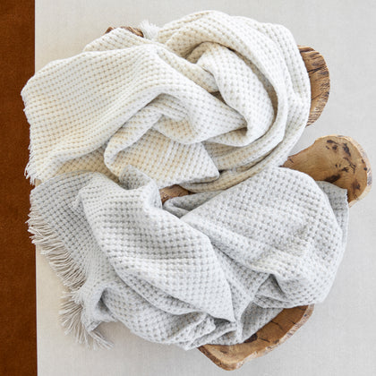 Overhead view of ivory and light gray Cleo knit throws with fringe, draped in a live-edge wood bowl.