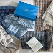 Overhead view of Cleo knit throws with fringe in navy, blue, ivory, tan, and charcoal, with a blue Matouk gift box and an open book.