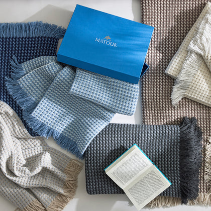 Overhead view of Cleo knit throws with fringe in navy, blue, ivory, tan, and charcoal, with a blue Matouk gift box and an open book.