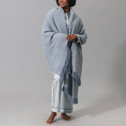 Model wearing light blue pajamas, draped in a blue and white Cleo knit throw with fringe.