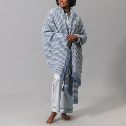 Model wearing light blue pajamas, draped in a blue and white Cleo knit throw with fringe.