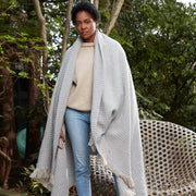 Model wrapped in a light gray Cleo knit throw with ivory fringe, standing outdoors in a wooded garden.