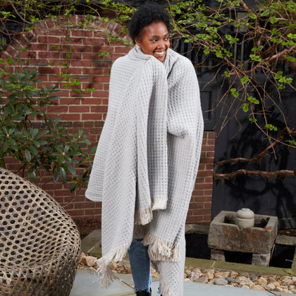 Model wrapped in a light gray Cleo knit throw with ivory fringe, smiling in an outdoor brick courtyard.