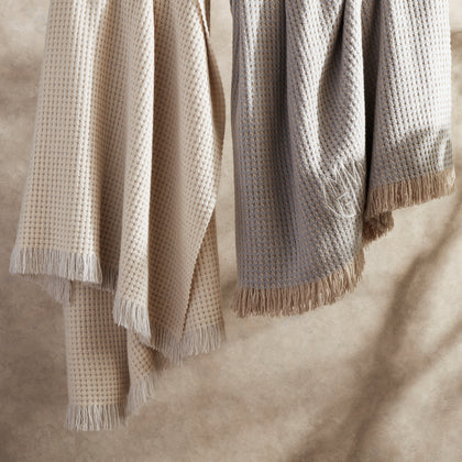 Ivory and gray Cleo knit throws with fringe and a monogram, draped over a bed.