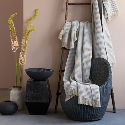 Light gray Cleo knit throw with ivory fringe, draped over a dark wicker chair and wood ladder in a styled vignette.