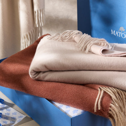 Fringed throws in tan and rust draped together, beside blue Matouk gift boxes.
