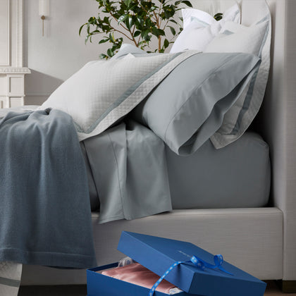 Bed with gray bedding and a quilted sham, gray throw, potted plant, open blue gift box with pink tissue paper on the floor.