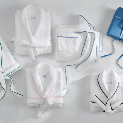 White terry robes with piped trim in green, blue, pink, and black, laid flat, beside a blue gift box.