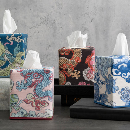 Four tissue box covers with dragon and cloud prints in navy, pink, brown, and blue-and-white, on books and concrete.