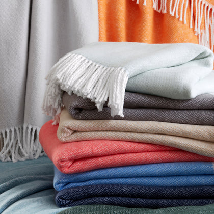 Pezzo fringed throws stacked in mint, gray, tan, coral, blue, navy, and sage, with orange and light gray throws draped behind.