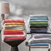 Pezzo fringed throws stacked in a full color range, spanning coral, yellow, blue, green, purple, and gray, on side tables and a bench.