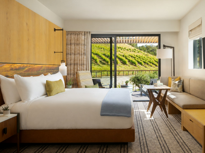 A modern luxury bedroom featuring a neatly made bed with white linens and a light blue throw, set against a large wood headboard. The room includes a built-in sofa, a small wooden table, and a large sliding glass door that offers a scenic view of a vineyard-covered green hillside.