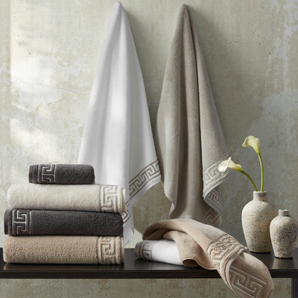 Adelphi towels with greek-key trim in white, tan, and charcoal, hung and stacked beside calla lilies.