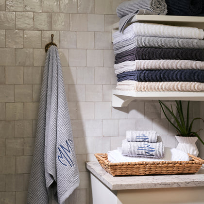 Gray monogrammed Aman towel hanging on a hook, with more stacked on a bathroom shelf.