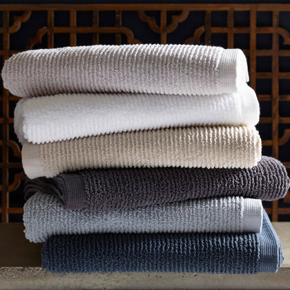 Ribbed Aman towels stacked in white, tan, gray, and navy, in front of a carved wood screen.