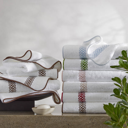 White Astor Braid towels with loop-embroidered trim in assorted colors, stacked beside soap and greenery.