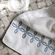 Close-up of a folded white towel with a blue-gray interlocking-loop embroidered border, on marble mosaic tile with a glass of water.