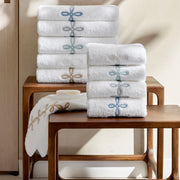 Stacks of folded white towels with interlocking-loop embroidered borders in blue, gray, and tan, on wood stools with a bar of soap.