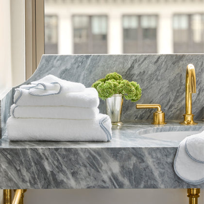 White towels with blue braided trim on a marble vanity with a gold faucet.