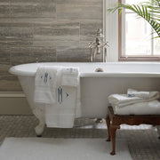 White Lotus towels with monogram embroidery, draped on a clawfoot tub and stacked on a wood stool.