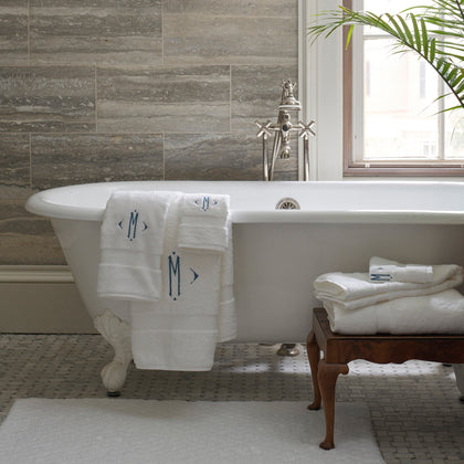 White Lotus towels with monogram embroidery, draped on a clawfoot tub and stacked on a wood stool.