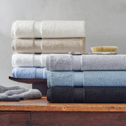 Lotus towels stacked in ivory, taupe, white, gray, pale blue, and black, on a wood dresser with a bath brush.