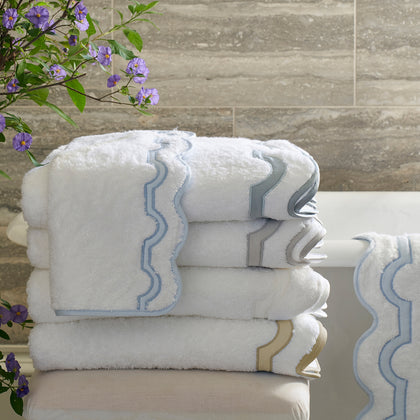 White Mirasol scalloped towels with wavy trim in blue, gray, and tan, stacked near purple flowers.