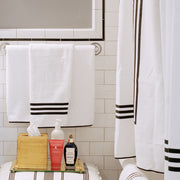 White bath towels with a black horizontal stripe border, white shower curtain with matching stripes, subway tile wall, bath products on a tray.