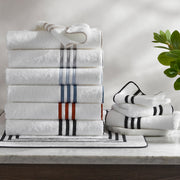Stacked folded white towels with striped borders in gray, blue, and red, black-striped bath mat and towels, marble counter, potted plant.