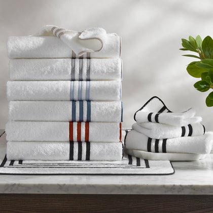Stacked folded white towels with striped borders in gray, blue, and red, black-striped bath mat and towels, marble counter, potted plant.