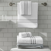White towels with a silver-gray horizontal stripe border, hanging on a wall hook and stacked, subway tile bathroom, gray bath mat.
