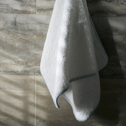 White Whipstitch towel with blue whipstitch trim, hanging against a marble wall.