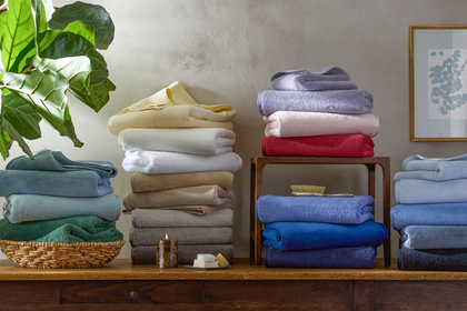 Stacks of Milagro bath towels in an assortment of colors, including sage green, cream, tan, lavender, blush, red, and blue.