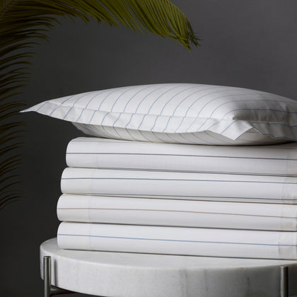 White Amalfi pinstripe sheets stacked on a marble table beside a palm frond.