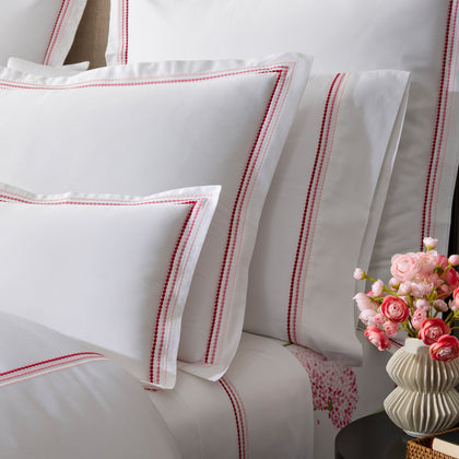 White pillow shams with triple-row embroidered dot borders in pink and red, a pink floral-print sheet, pink ranunculus in a vase.