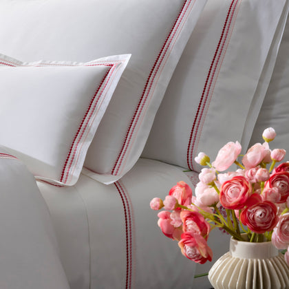 Close-up of white pillow shams with triple-row embroidered dot borders in pink and red, pink ranunculus flowers in a vase.
