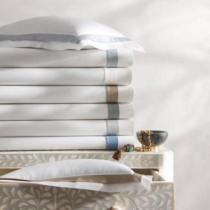 White Ambrose sheets with satin bordered trim in blue and tan, stacked on an inlaid dresser.