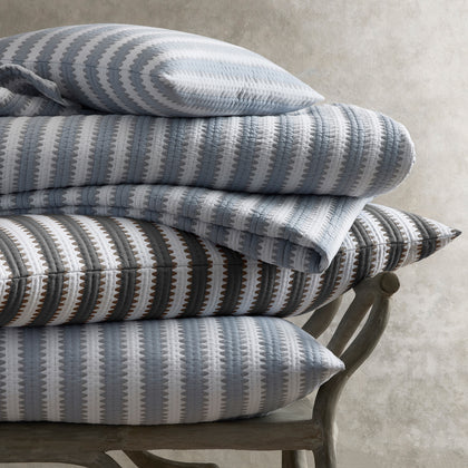 Apollo Stripe Matelasse quilted bedding in blue and gray, stacked on a wooden bench.