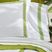 Close-up of white AstorBraid pillow shams and sheets with green looped-braid embroidered borders, against a botanical-print pillow.
