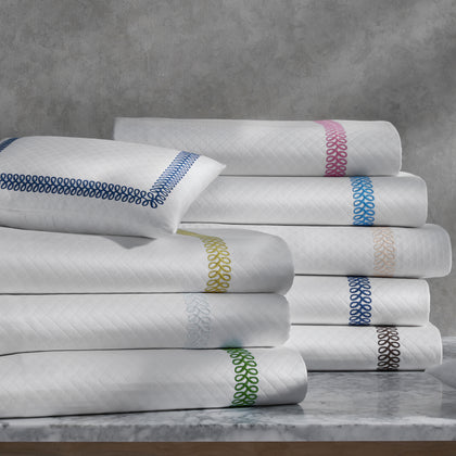 White Astor Braid quilted bedding with loop-embroidered trim in a rainbow of colors, stacked on a marble table.
