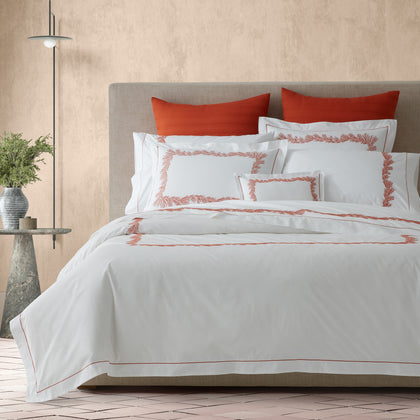 White Atoll bedding with coral embroidered border, styled on a bed with rust-colored pillows.