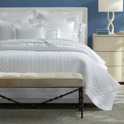 White Ava quilted bedding with blue floral embroidered trim, styled on a bed against a blue wall.